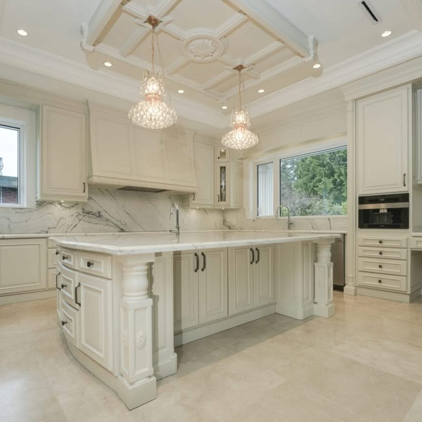 Bright luxury white kitchen with a large central island, marble surfaces, pendant lights, custom cabinets and a decorative tray ceiling.
