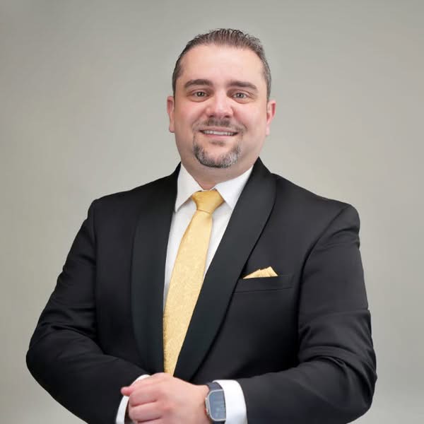 Smiling man in a dark suit and yellow tie standing with his hands together against a neutral background
