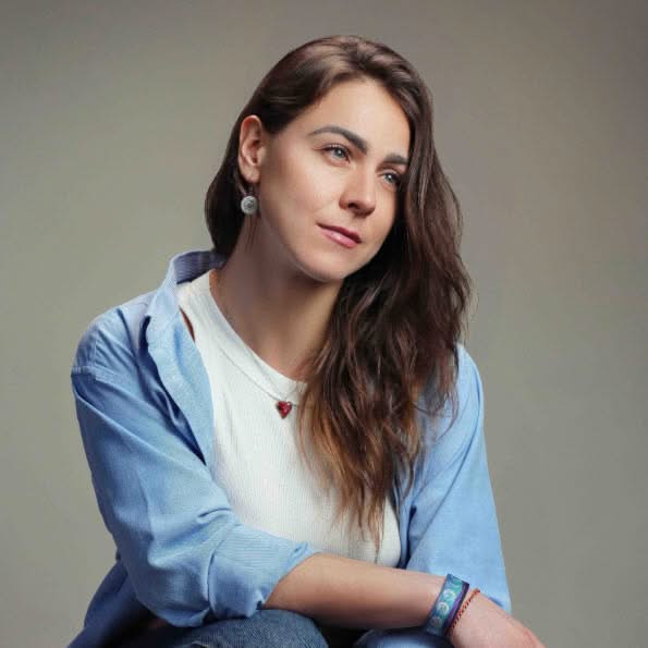 Portrait of a smiling woman with long brown hair wearing a blue jacket and facing the camera against a neutral background.