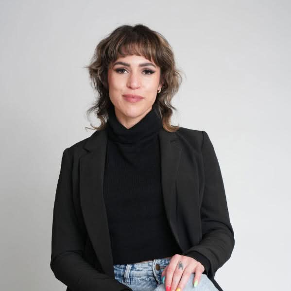 Woman with short brown hair seated in a black suit and turtleneck against a light grey studio background