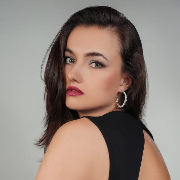 Brunette woman wearing a black sleeveless outfit looking over her shoulder with red lipstick and silver hoop earrings.