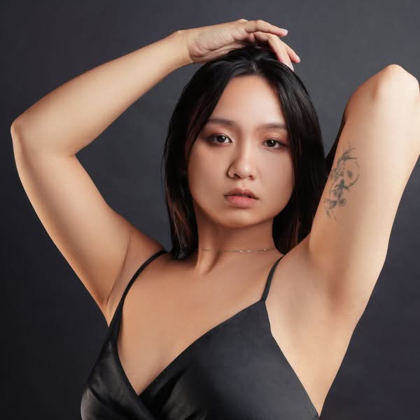 Dark haired woman wearing a black dress and posing with both arms raised against a charcoal studio background