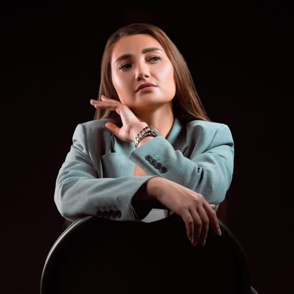 Woman wearing a light blue jacket seated against a black background with her hand raised in an expressive studio portrait