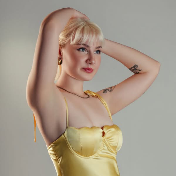 Blonde woman wearing a gold strapless dress and posing with her arms raised against a light studio background