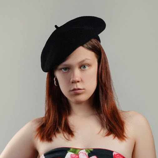 Professional headshot of a serious red haired woman wearing a black beret and strapless black outfit against a light studio background