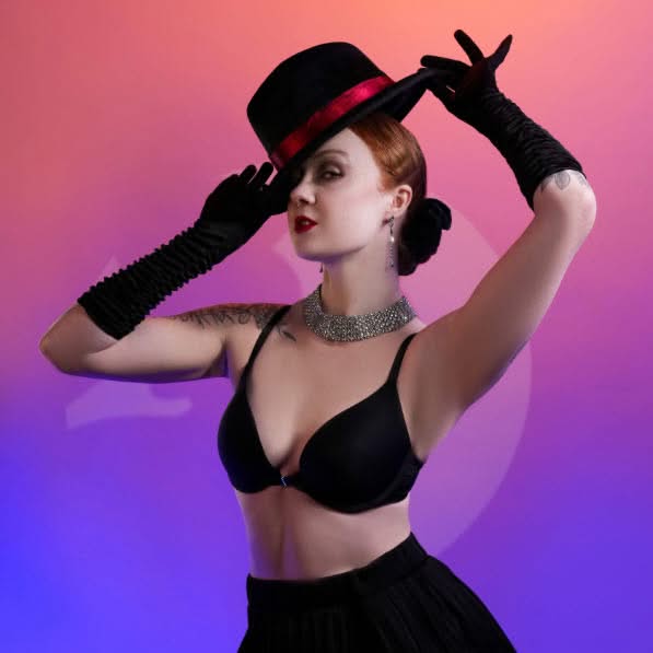 Woman wearing a black glamour outfit, long gloves and a tilted hat while posing against pink and purple lighting