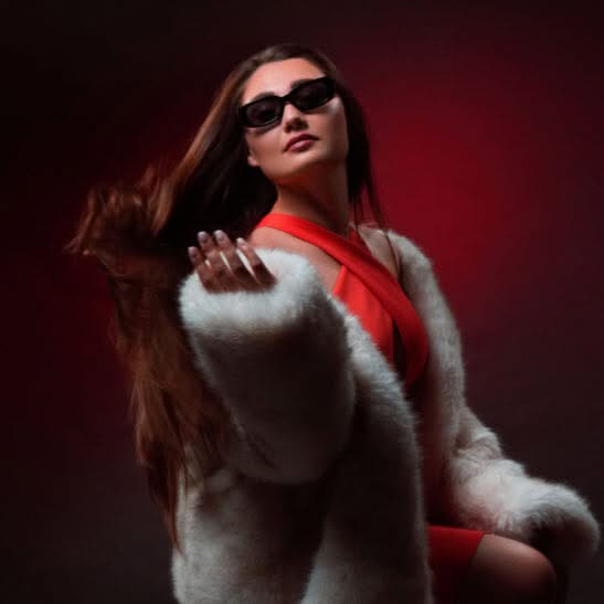 Woman wearing sunglasses and a light fitted outfit posing under dramatic red studio lighting
