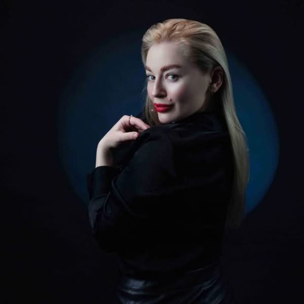 Blonde woman in black clothing photographed in profile with dramatic blue rim lighting against a dark background