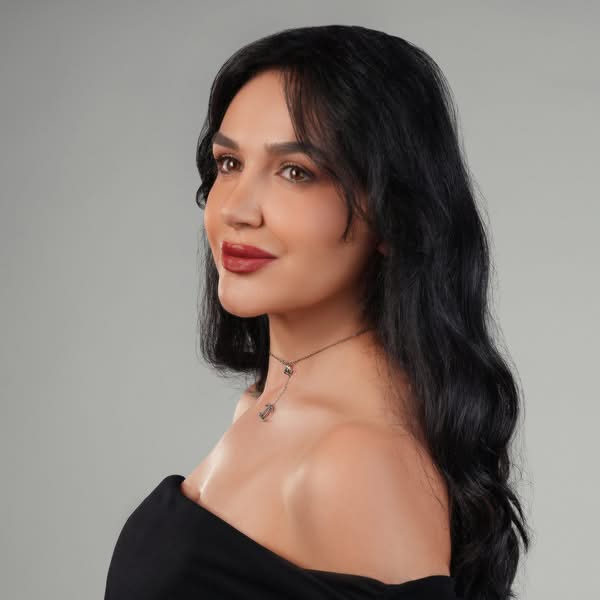 Woman with long black hair wearing a black off shoulder top and posing at an angle against a grey background