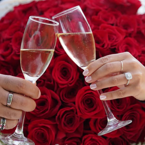 Couple holding champagne glasses above red roses with engagement rings visible on their hands