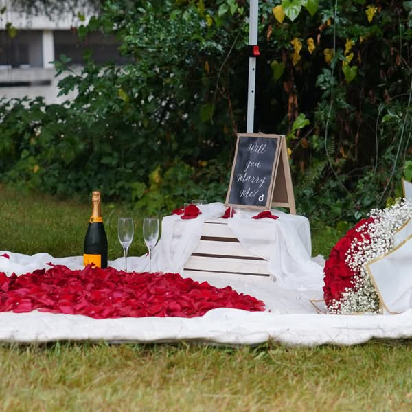 Romantic outdoor proposal table decorated with red rose petals, champagne and a white cake