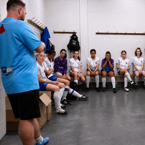 Coach in a blue shirt speaking to soccer players seated on locker room benches in white uniforms