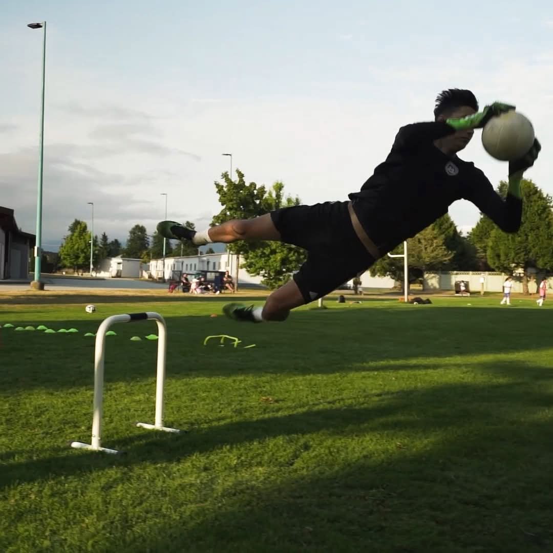 Soccer goalkeeper in a dark uniform diving sideways to catch the ball during an outdoor training session